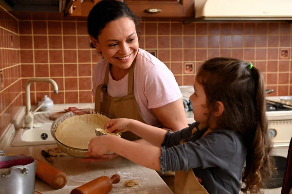 Beautiful woman, a caring mom and her lovely daughter smiling, laughing, playing, enjoying cooking together in kitchen, holding molds with rolled out dough, preparing homemade tartlets and cherry pie