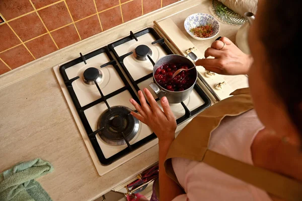 Overhead view of pretty woman, a housewife in a beige chefs apron, standing by the stove in home kitchen and drizzling sugar with cherries while preparing caramelized filling for homemade festive pie