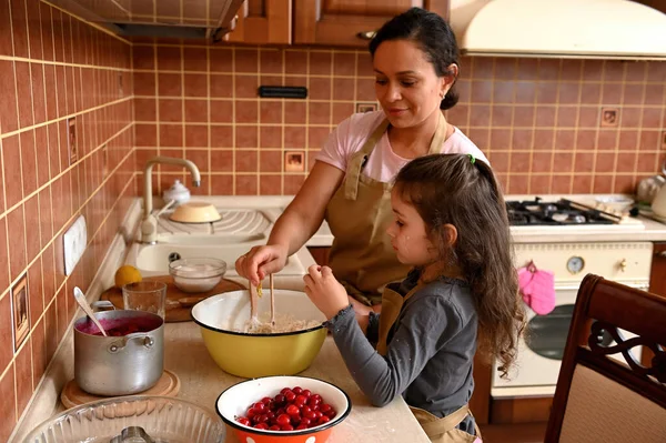 Happy multi-ethnic family, a loving mother and daughter, both wearing beige chefs aprons, baking pastries together, kneading dough for homemade festive cherry pie in the home kitchen. Family look