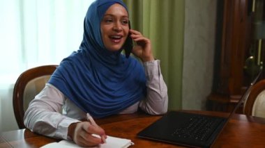 Kafası tesettüre bulanmış, cep telefonuyla konuşan güzel bir Arap kadın, lüks ofis içerisinden dizüstü bilgisayarla çalışırken notlar alıyor. Kavram: Müslüman kadınlar için iş fırsatları