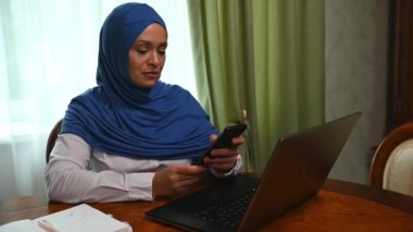 Mavi tesettürlü güzel, şık, Müslüman bir kadın iş ortaklarına çağrı yapıyor, bir dizüstü bilgisayarın önünde ahşap masada oturuyor ve merkez ofisteki yeni bir proje üzerinde çalışıyor.