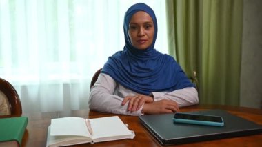 Orta Doğu kökenli çekici Müslüman bir kadının kendinden emin portresi, mavi başörtüsü takmış, kameraya özgüvenle bakıyor, toplantı odasında dizüstü bilgisayar ve akıllı telefonla ahşap bir masada oturuyor.