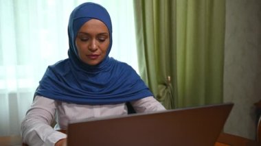 Kafası tesettüre bulanmış, tahta masada oturan ve dizüstü bilgisayarla yeni bir proje üzerinde çalışan kendinden emin, Ortadoğulu Müslüman güzel bir kadın. Planlama işi. Çevrimiçi iletişim. Serbest meslek