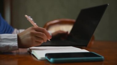 Yakın plan. Kalem, yazı ve dizüstü bilgisayar üzerinde çalışırken not alan kadınlara özen göster. Cep telefonu ahşap masada. İş, proje, çevrimiçi uzaktan çalışma