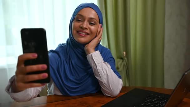 ヒジャーブの自然美のムスリム女性 スマートフォンを使用して 自撮りを取ります 彼女のブログのためにビデオを記録します ビデオリンクで呼び出しをしながら 彼女の対話者を迎えます 美しいおもちゃの笑顔を笑顔 — ストック動画