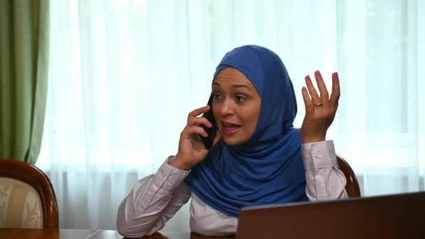 電話での会話の失敗の間に不安を感じながら 自宅のオフィスで木製の机に座って 携帯電話で話すビジネス女性 成功した起業家 ヒジャーブのイスラム教徒の女性をマルチタスク — ストック動画