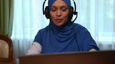 Yakın plan Müslüman, tesettürlü çekici bir kadın, kulaklık takıyor, evden işe gidiyor, dizüstü bilgisayar kullanıyor, iş ortakları veya müşterileriyle video bağlantısı üzerinden konuşuyor, çevrimiçi bir konferans sırasında