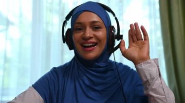 Laptop monitörüne eklenecek video: Başörtüsü ve ses kulaklığı takan güzel Müslüman kadının ön görünümü, bir video konferansı sırasında veya öğrencilerle uzaktan çevrimiçi ders sırasında kameraya bakar
