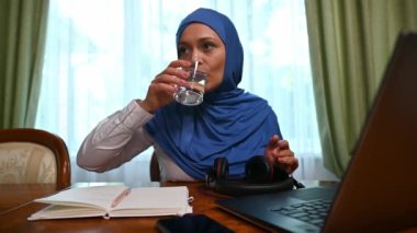Başörtülü güzel İslamcı kadın, mikrofon ve kulaklıkla konuşmadan önce bir yudum su alıyor, çağrı merkezinde çalışıyor. Uzaktan kumanda. Çevrimiçi ders kulaklığı. Video konferansı