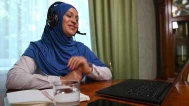 Güzel başarılı tesettürlü Müslüman bir kadın, ev ofisinin ahşap masasında oturuyor, internetteki bir video konferansı sırasında dizüstü bilgisayar ve kulaklık kullanıyor. Sosyal mesafe. Uzak meslek