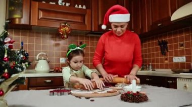 Neşeli çoklu etnik kadın, kırmızı kazaklı ve Noel Baba şapkalı sevgi dolu anne, merdane kullanıyor, zencefilli kurabiye için hamur atıyor ve tatlı kızının kurabiye kalıplarıyla kalıp kesmesine yardım ediyor.