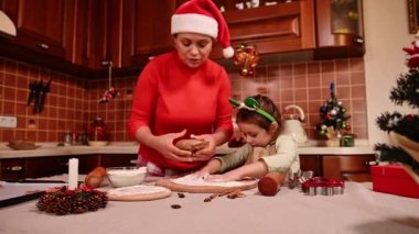 Sevimli koyu saçlı kadın, şefkatli anne ve tatlı kızı Noel karnavalında aşınıyor, hamur yoğuruyor ve yuvarlanıyor, ev mutfağında zencefilli kurabiye pişiriyorlar.