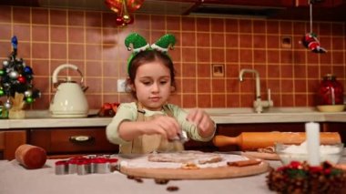 Kafasında elf halkası ve bej rengi önlüklü sevimli beyaz kız, mutfak adasında duruyor ve zencefilli ekmek hamurundan kalıp kesiyor, Noel için lezzetli kurabiyeler hazırlıyor.