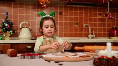 Sevimli beyaz çocuk, elf çemberi ve şef önlüğü olan küçük bir kız zencefilli ekmek hamuru kalıplarını kesiyor, kameraya gösteriyor, ev mutfağında kurabiye pişirmenin keyfini çıkarıyor. Noel neşesi