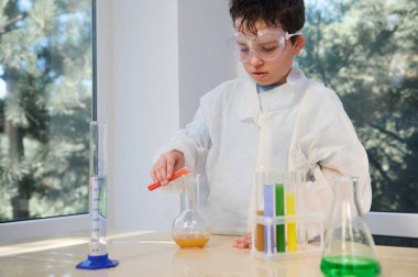 Beyaz önlüklü ve güvenlik gözlüklü ilkokul öğrencisi, kimyasal deney yapıyor, deney tüpünden laboratuvar tüpüne sıvı döküyor, fen dersinde kimya öğreniyor. Eğitim konsepti.