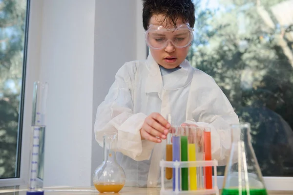 Confident smart Caucasian boy wearing safety goggles and white lab coat, learning Chemistry in the school science laboratory, conducting chemical experiment, using regents, chemicals and glass labware