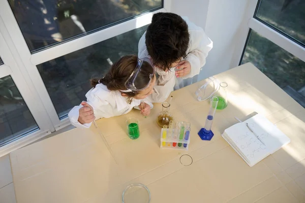 Overhead view of school kids, wearing safety goggles and white lab coat, learning Chemistry in the school science laboratory, conducting chemical experiment, using regents, chemicals and glass labware