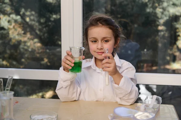 Beautiful little girl chemist conducts a chemical reaction, pours a solution of soda into citric acid, observes the chemical reaction taking place in test tube, studies Chemistry in school laboratory