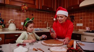 Mutlu Noeller ve mutlu kış tatilleri. Aile hazırlığı bayram yemeği. Kırmızı kazaklı ve Noel Baba şapkalı neşeli bir anne, elf çemberli şirin bir kız, ev yapımı zencefilli kurabiye pişiriyor.