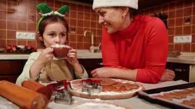 Noel Baba şapkalı güzel bir kadın, sevgi dolu bir anne, sevimli kızına hayran, elf çemberli küçük bir kız, ev mutfağında marşmelovla sıcak çikolata içerken, Noel için zencefilli kurabiye pişiriyor.