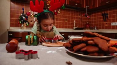 Beyaz tenli, bej bir önlük ve geyik boynuzu çemberi takmış, kurabiye kesiciyle zencefilli hamurdan şekiller kesmeye odaklanmış afacan küçük bir kız. Güzel bir Noel arifesinde yemek pişirmek.