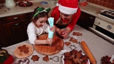 Noel Baba şapkalı mutlu bir anne, kızına bir şeyler öğretiyor. Elf çemberli küçük bir kız, pasta torbasında krema kullanıyor, ev mutfağında zencefilli kurabiye süslüyor. Noel zamanı. - Aile. Yemek pişirmek. Kış tatili