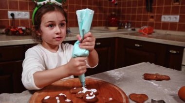Güzel bir çocuk, beyaz süveter ve elf potası giymiş sevimli beyaz bir kız fırında pişmiş zencefilli kurabiyelerin üzerindeki kremadan krema sıkıyor. Çocuk Noel için süsleme hamur işlerini cila ile öğreniyor.