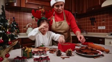 Mutlu anne ve kız, Noel Baba şapkası ve geyik boynuzu çemberiyle keten ip kullanıyor, taze zencefilli kurabiye bağlıyor, onları hediye kutusuna koyuyor, Noel atmosferinde eğlencenin tadını çıkarıyor.