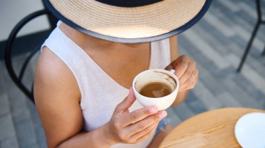 Hasır şapkalı ve yaz elbisesi giymiş, elinde beyaz bir fincan kahve Amerikano 'su olan, sıcak bir yaz terasında kahve molası veren, tanınmayan gizemli bir kadının tepeden görünüşü.