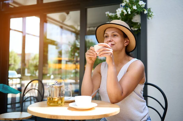 Relaxed beautiful dark-haired multiethnic woman in a straw hat and summer dress, sitting at a table outdoors in cozy cafe, enjoying a coffee break on sunny day. Lifestye. Recreation. Chill