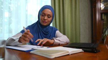 Güzel, kendine güvenen Orta Doğulu, kafası mavi tesettüre bulanmış güzel bir kadın. Ev ofisinden laptopla bilgisayarla seyahat ederken not defterine not alıyor. Modern Müslüman kadınlar için çalışma fırsatları