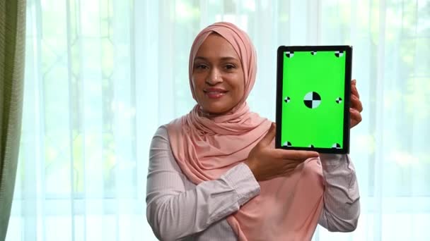 漂亮的阿拉伯穆斯林妇女戴着粉色头巾 手持带有绿色彩色按键屏幕的数码平板电脑 微笑着看着相机 插入促销视频剪辑 电影或广告文本的理想模型 — 图库视频影像