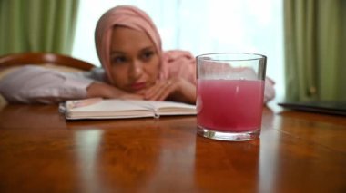 Pembe tesettürlü Arap Müslüman kadın, bir bardak suda eriyen parlak bir tableti izliyor. Çoklu görev yapan kadın, dizüstü bilgisayardaki aşırı çalışmadan kaynaklanan gerilimden dolayı migreni yatıştırmak için ağrı kesici alır.