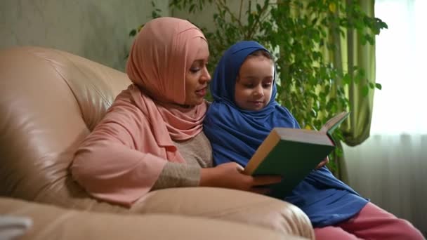 ピンクのヒジャーブの美しいイスラム教徒の女性 彼女の愛らしい小さな娘におとぎ話を読んで 居心地の良い家のインテリアの快適なアームチェアに一緒に座って — ストック動画