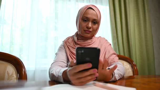ピンクのヒジャーブの魅力的な中東イスラム教徒の女性 自宅でリラックスしながらスマートフォンを使用して ショックを感じます ストレスと失望 彼女の携帯電話で悪いニュースを読んで 恐怖と悲しみを感じます — ストック動画