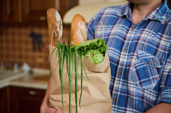 Cropped view of an eco paper shopping bag with loaves of whole grain bread, healthy vegan food in mans hands before unpacking in the home kitchen. Healthy eating and lifestyle. Veganism