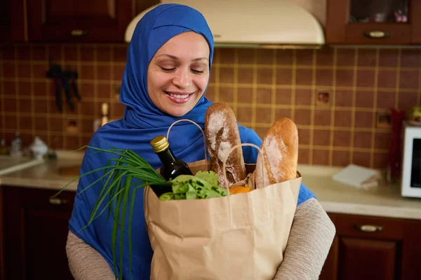 Healthy purchasing from grocery. Food delivery. Beautiful Middle-Eastern Muslim woman wearing a blue hijab, unpacking a shopping bag with healthy food, standing against cozy home kitchen background