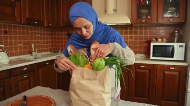 Mavi tesettürlü güzel bir Orta Doğulu Müslüman kadın, alışveriş ekosistem kâğıdından bir poşet açıyor ve evdeki mutfakta erzakla birlikte. Sağlıklı yiyecekler. Vejetaryenlik. Sağlıklı bir yaşam tarzı. Ev hayatı.