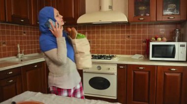 Büyüleyici orta yaşlı, modern Arap kadın mavi tesettüre ve günlük kıyafetlere sahip, cep telefonuyla konuşan, sıcak ev mutfağındaki masaya organik yiyecekler koyan.