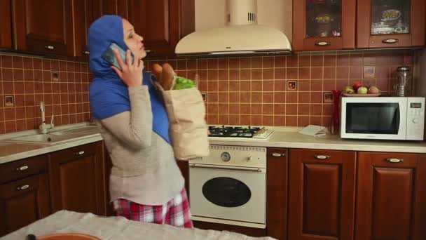 迷人的中年妇女 穿着蓝色头巾和休闲装 一边用手机聊天 一边把装有有机食品的购物袋放在舒适的家庭厨房桌上 — 图库视频影像