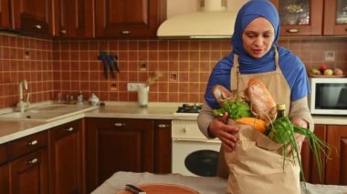 Gülümseyen Müslüman bir kadın market alışverişinden sonra organik sebzeler ve meyvelerle dolu poşetleri açıyor. Mutlu Orta Doğulu ev hanımı, evin mutfağında duruyor. Boşluğu kopyala