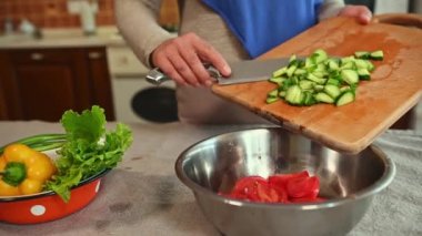 Tanınmayan bir ev kadınının yakın çekimi. Tahtadan kesilmiş salatalıkları metal bir kaseye koyup organik mevsimlik taze sebzelerden çiğ vegan salatası pişiriyor.