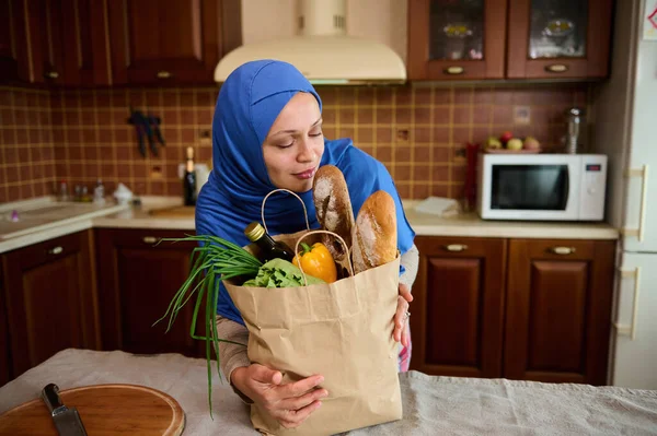 Muslim Arab pretty woman with head covered in blue hijab, enjoys the aroma of a loaf of freshly baked healthy bread in a grocery bag. Housewife arriving home after shopping for organic healthy food