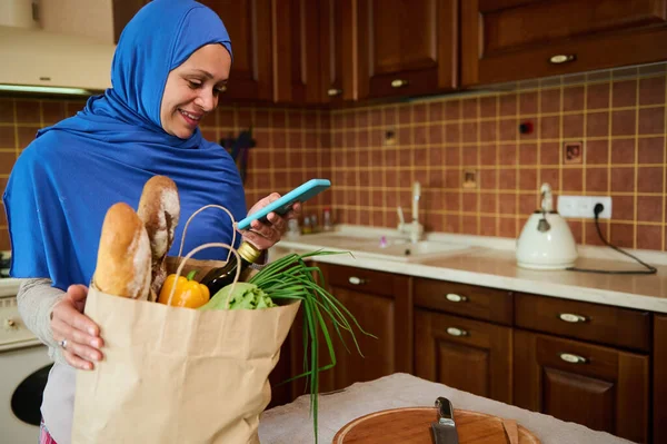 Charming Arab Muslim woman with head covered in hijab using mobile phone, checking expenses before unpacking eco bag with organic vegetables and healthy food, while arriving home from grocery shopping