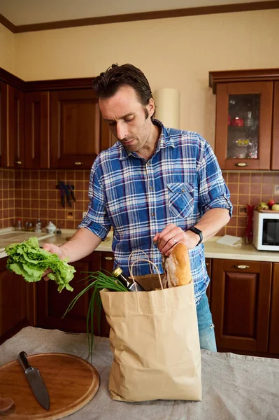 Handsome Middle-aged Caucasian man sorting organic fresh vegetables while unpacking grocery shopping bag in the kitchen at home. Food delivery. Healthy grocery purchasing in farmers markets and stores