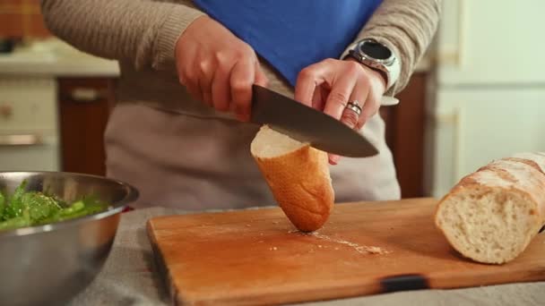 木製のまな板の上に2つの半分に全粒粉パンローフを切断女性の作物のビュー 夕食のためにパンを切り 自宅のキッチンテーブルに立つ女性 — ストック動画