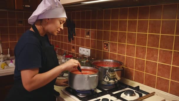 白いコック帽と黒エプロンの多民族の女性 ストーブのそばに立つ 自宅でキッチンで熟した有機ジューシーなトマトから自家製パスタを準備しながら 沸騰トマトジュースを攪拌 — ストック動画