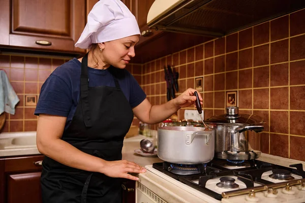 Multiethnic woman in white chefs cap and black apron, stands by the stove, stirring boiling tomato juice while preparing homemade passata from ripe organic juicy tomatoes in the kitchen at home