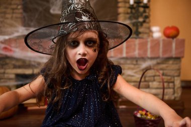 Yanağında örümcek resmi olan, büyücü şapkası ve cadı elbisesi giyen gotik küçük bir kızın yakın plan portresi büyüler yapıyor ve kameraya bakıyor. Cadılar Bayramı partisi. Enchantress. Büyücü.