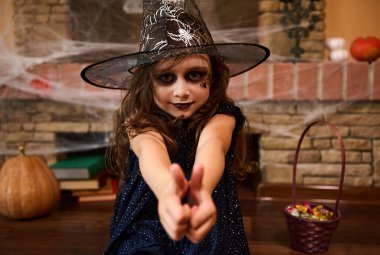 Büyücü şapkalı gotik küçük cadı kız Cadılar Bayramı ziyafetleriyle çevrili bir şekilde örümcek ağıyla kaplı şöminenin karşısında oturuyor ve kameraya baş parmağını gösteriyor. Kitapları yerde yatarken büyülüyor. Cadılar Bayramı partisi
